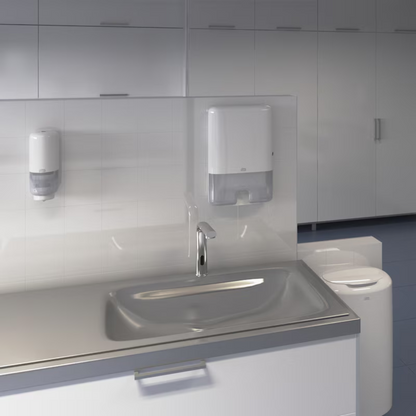 Gezeigt wird ein modernes Waschbecken in einer öffentlichen Toilette mit automatischem Wasserhahn. Über dem Waschbecken sind ein Tork Xpress® 552000 Spender für Multifold Handtücher H2 von Essity Professional Hygiene Germany GmbH und ein Seifenspender an einer gefliesten Wand montiert. Im Hintergrund ist eine Reihe weißer Schränke zu sehen.