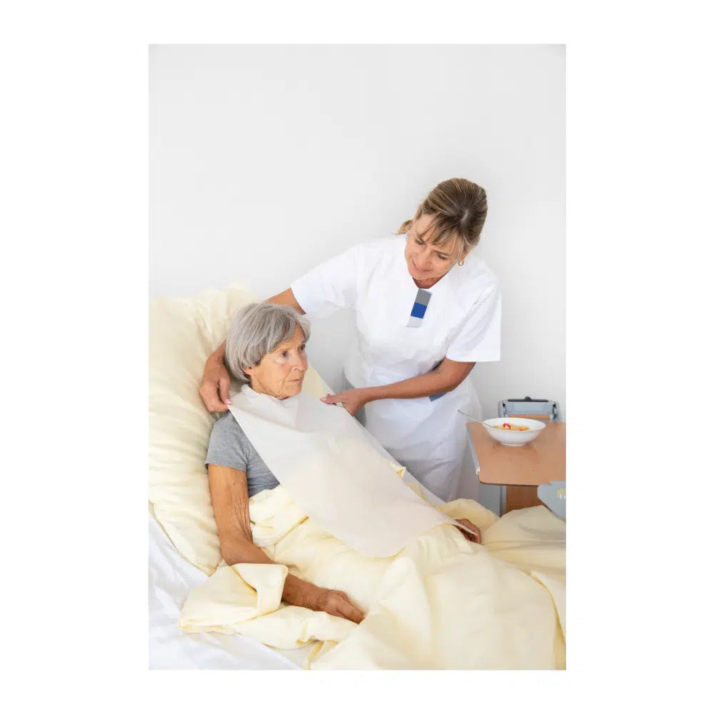 Eine Krankenschwester richtet die Decke für eine ältere Frau, die aufrecht in einem Krankenhausbett sitzt. Beide wirken aufmerksam und konzentriert. Ein Tablett mit einer Schüssel und Vala® Fit Tape Einmal-Kleidungsschutz ist zu sehen.