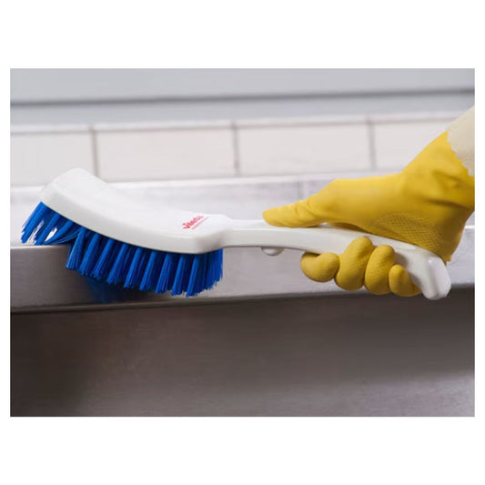 Eine Nahaufnahme einer Hand mit einem gelben Gummihandschuh, die eine weiß geflieste Oberfläche mit einer Vileda Professional Stielbürste mit kurzem Griff blau – 30 c schrubbt| Packung (1 Stück) mit hochwertigen Borsten.