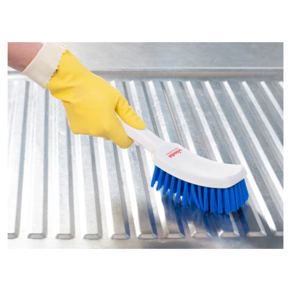 Eine Person mit einem gelben Gummihandschuh reinigt eine Metalloberfläche mit einer Vileda Professional Stielbürste mit kurzem Griff blau. Die Umgebung lässt vermuten, dass der Fokus auf gründlicher, hygienischer Reinigung liegt.