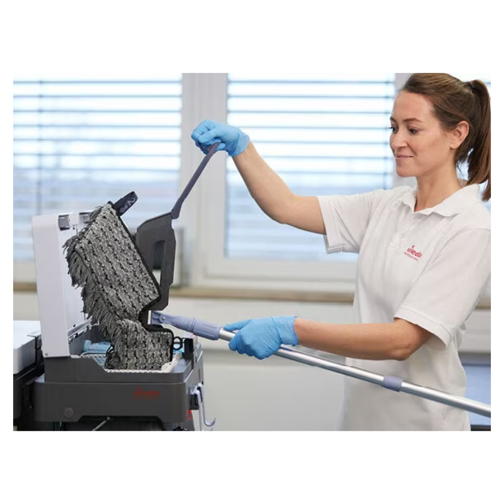 Eine Frau im Laborkittel und mit Handschuhen bedient eine Maschine in einer Laborumgebung und führt einen Vileda Professional Swep Duo r-MicroCombi Mop - 50 cm | Packung (1 Stück) mit langem Griff in das Gerät ein. Sie scheint sich auf ihre Aufgabe zu konzentrieren.