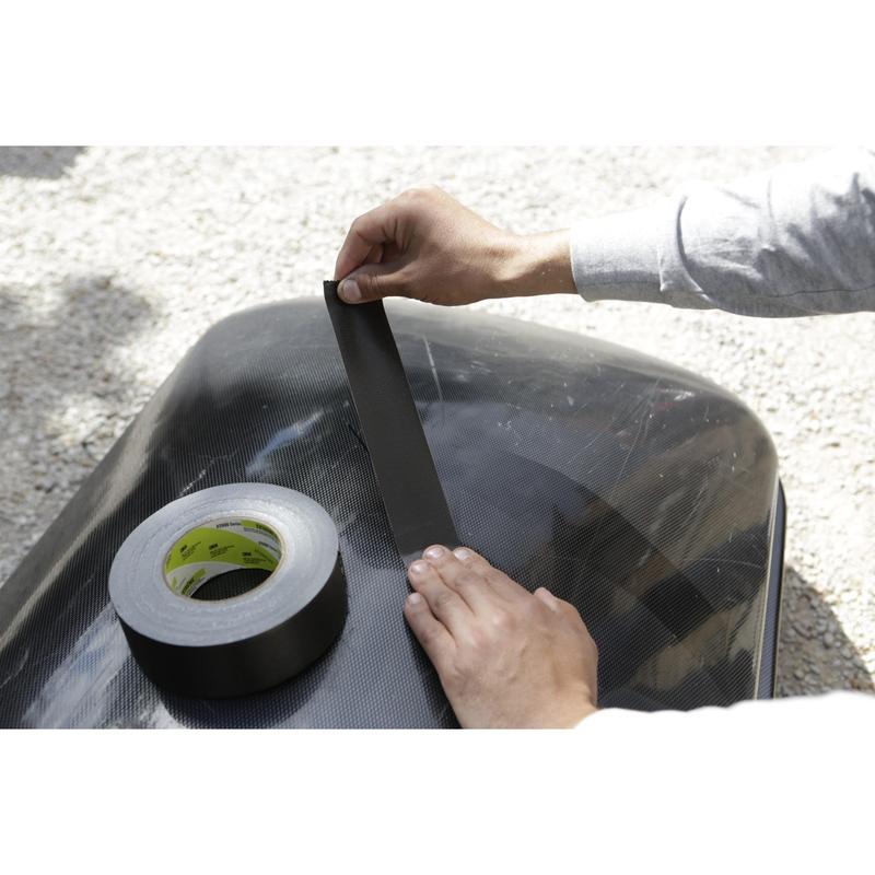 Someone is applying a strip of Scotch® Extremium™ Ultra High Performance Tape DT17 (25 m x 48 mm) from 3M Deutschland GmbH to a smooth, dark exterior surface. Nearby lies a roll of this high-performance tape; gravel can be seen in the blurred background.
