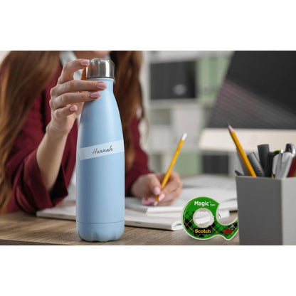 A person holds a light blue water bottle labeled "Hannah" at a desk with notebooks, a pencil, a 19 mm x 25 m Scotch® Magic™ Invisible Tape refill pack from 3M Deutschland GmbH, and a pen holder in an office or study.