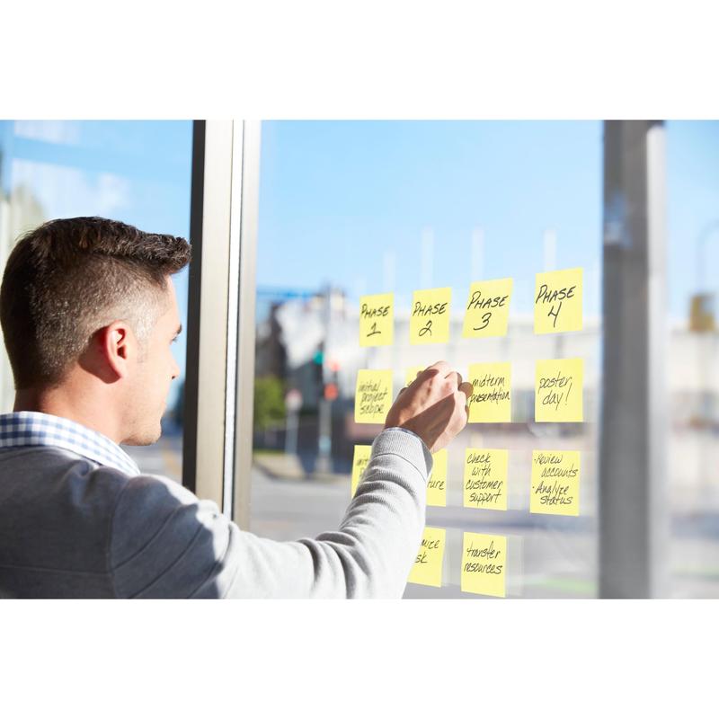 A person uses Post-it® Super Sticky Notes (76 x 76 mm, 90 sheets, PEFC-certified) from 3M Deutschland GmbH to organize project phases on a glass window where sunlight falls in and a street can be seen.