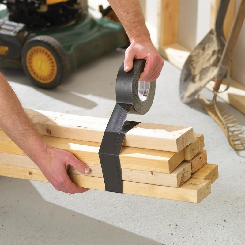 In a garage, someone is wrapping the Scotch® Extremium™ Ultra High Performance Tape DT17 (25 m x 48 mm) from 3M Deutschland GmbH around wooden boards, while a lawnmower, a rake, and a shovel can be seen in the background.