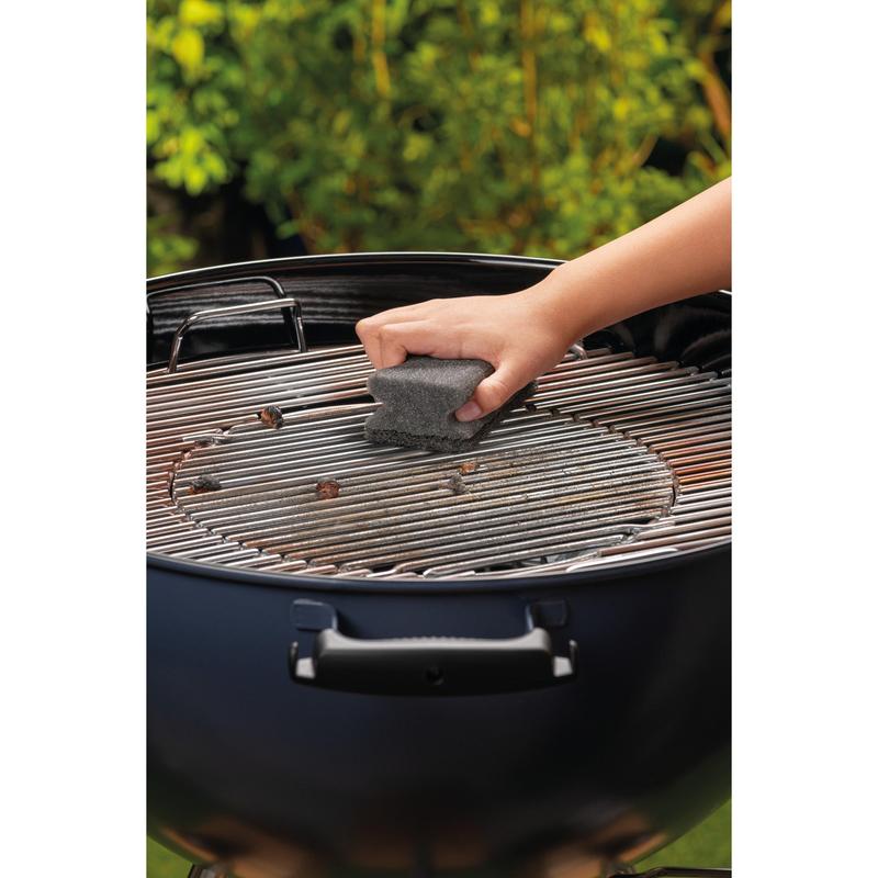 Eine Hand reinigt den Metallgrill eines runden Holzkohlegrills mit einem Scotch-Brite® Grillgriffschwamm (1 Stück pro Packung) der 3M Deutschland GmbH. Grünes Laub ist im Hintergrund unscharf.