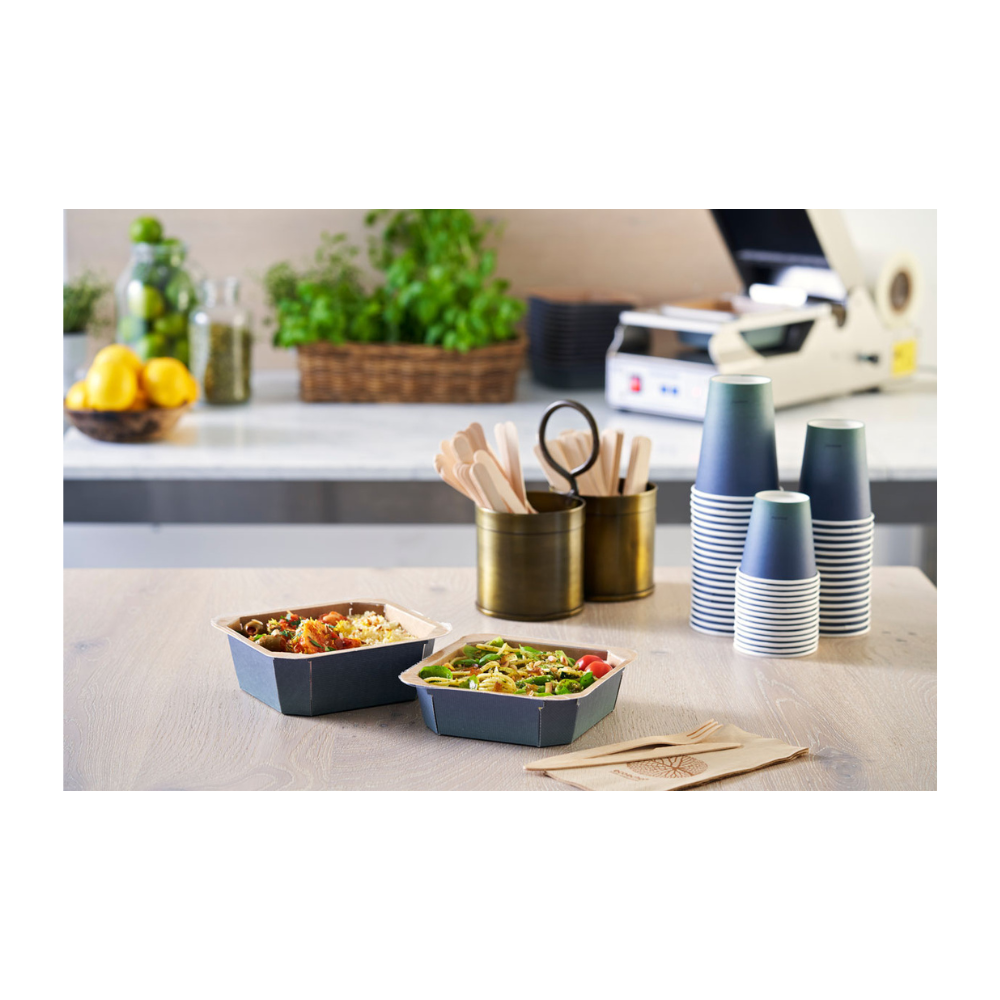 On the table are two take-away bowls, flanked by stacked Duni Urban Coffee Cups PAP/PLA from Duni GmbH, wooden cutlery in a holder, and napkins. Lemons, glasses, and herbs in the background contribute to the sustainable ambiance of this food preparation area.